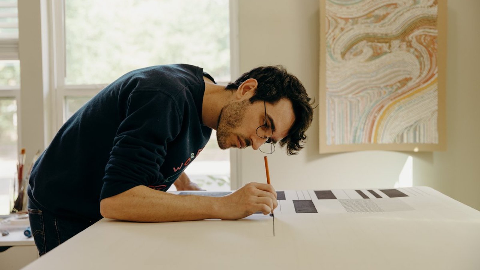 Tyler Hobbs bent over a table drawing on paper, a framed work with flowing colored bands behind him